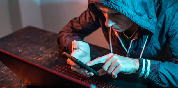 istock Hacker with phone is typing on a laptop keyboard in a dark room under a neon light. Cybercrime fraud and identity theft 1313967074