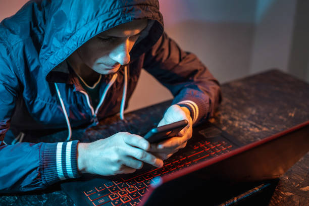 istock Hacker with phone is typing on a laptop keyboard in a dark room under a neon light. Cybercrime fraud and identity theft 1313967080