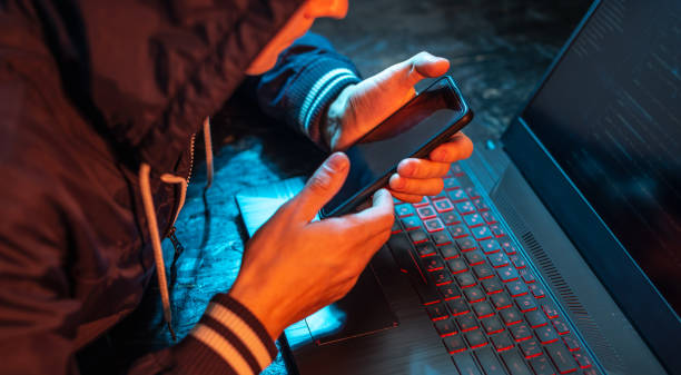 istock Hacker with phone is typing on a laptop keyboard in a dark room under a neon light. Cybercrime fraud and identity theft 1313967100