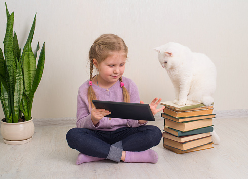 Little smiling girl sitting on the floor with her cat and tablet computer. Education leisure pets technology concept.