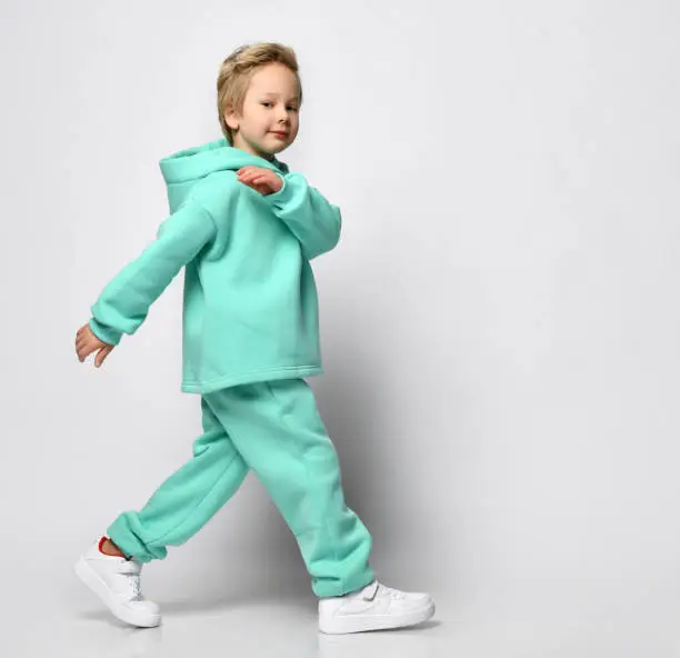 Full portrait of stylish little boy in warm tracksuit against gray background. A boy in a turquoise fleece suit and sneakers stands sideways to the camera near free space for text.