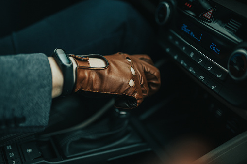 Close-up on male hand gripping gearstick of a manual car with brown leather driver gloves on and black smart watch on right hand