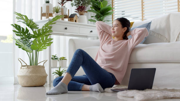 young attractive beautiful asia female girl or university student sit smile look outside window put arm hand back behind head at sofa couch living room feeling relax comfort at cozy home houseplant. - lying down women laptop freedom imagens e fotografias de stock