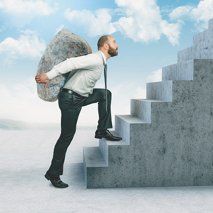 businessman climbs the stairs with a large stone on his back.