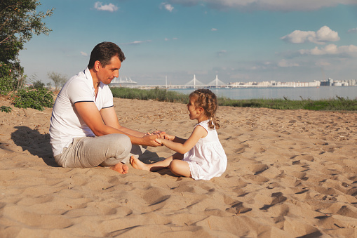 dad and little daughter in white dress are sitting on beach and play communicate and hold hands, copy space