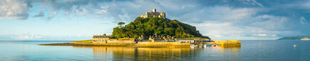 Sunlit island in tranquil sea St Michaels Mount panorama Cornwall Gentle ocean tide ebbing across the iconic cobbled causeway as warm summer sunlight illuminates the historic chapel, hamlet and harbour of St. Michael's Mount, Marazion, Cornwall, UK. marazion photos stock pictures, royalty-free photos & images