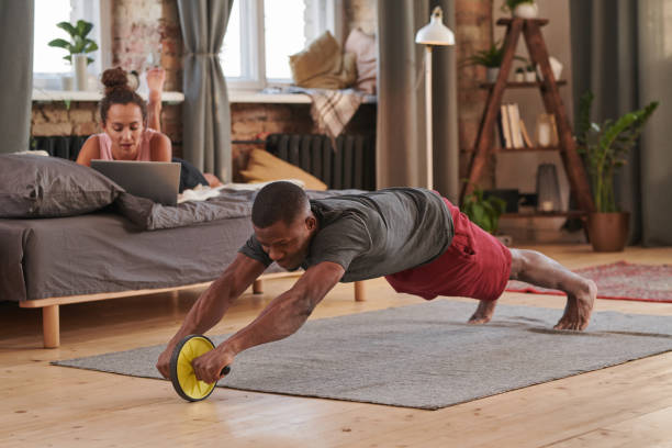 リビングルームの床にabローラーを使用して強化運動をしている現代のyoun大人アフリカ系アメリカ人男性の水平ショット - 腹筋ローラー ストックフォトと画像
