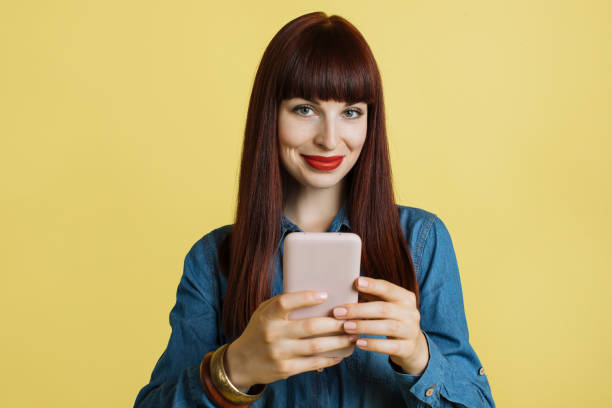 istock Using mobile phone, typing sms message. People and technologies. Close up of smiling young woman in jeans shirt posing isolated on pastel yellow background and using smartphone 1313998847