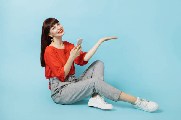 istock Young lady in smart casual wear, holding phone, showing imaginary something, empty copy space for some text, product or slogan, on the open hand palm, sitting isolated on blue background. 1314000147