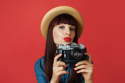 Hobby, taking photos, travel concept. Gorgeous young woman in hat, photographer amateur, smiling and making air kiss, taking picture on old vintage camera, on colorful red studio background