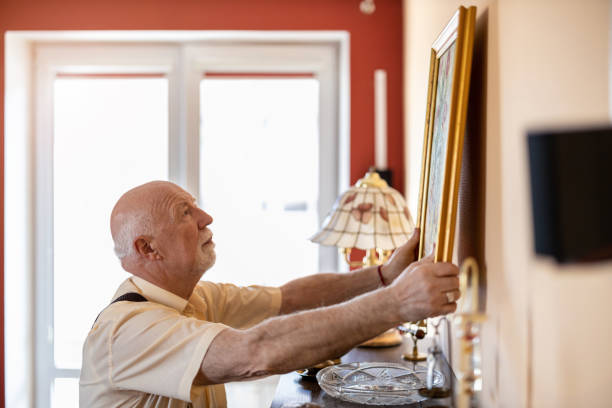 hombre mayor colocando una pintura en la pared en su casa - pintura producto artístico fotos fotografías e imágenes de stock