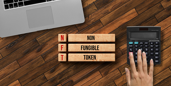 wooden blocks with message NFT - NON FUNGIBLE TOKEN and a hand over a calculator on wooden background