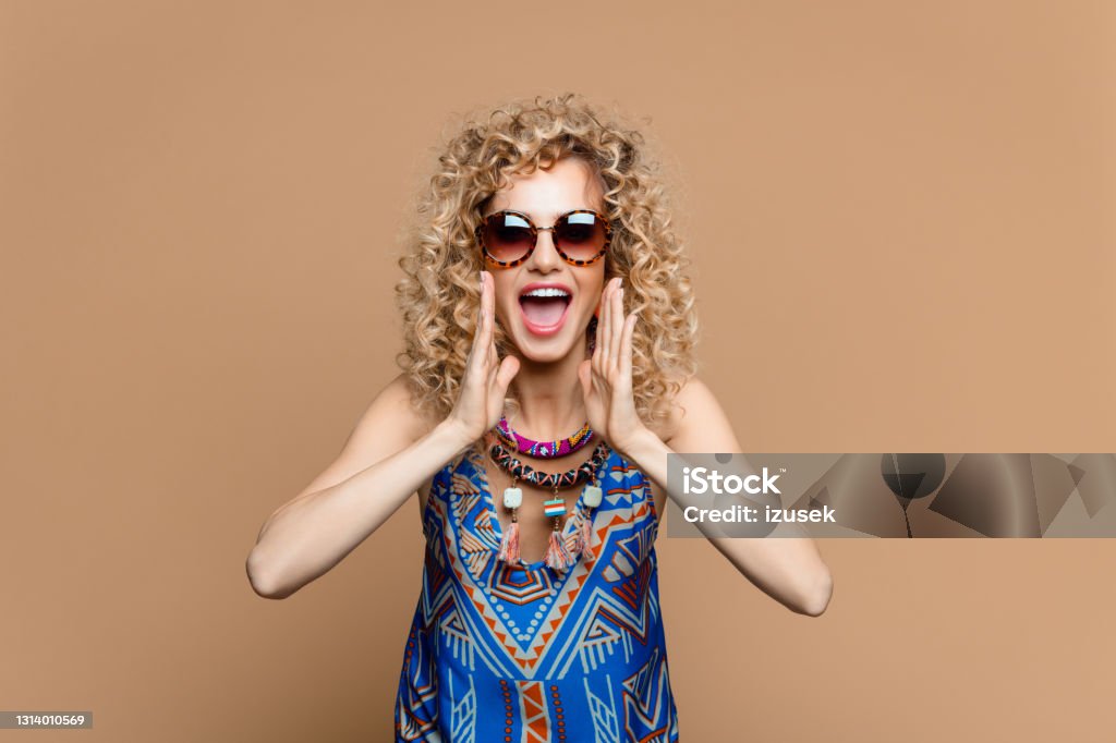 Excited woman in boho style outfit against brown background Summer portrait of beautiful long curly blond hair young woman wearing boho style dress and jewelry, shouting at camera. Studio shot on brown background. Summer Stock Photo