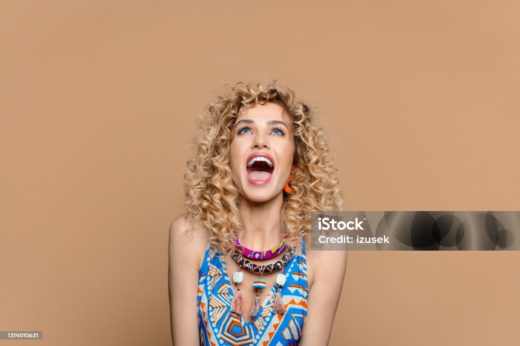 Excited woman in boho style outfit against brown background Summer portrait of beautiful long curly blond hair young woman wearing boho style dress and jewelry, looking up and laughing. Studio shot on brown background. 25-29 Years Stock Photo