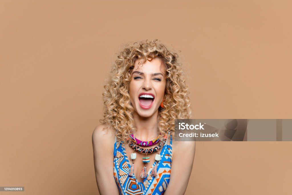 Excited woman in boho style outfit against brown background Summer portrait of beautiful long curly blond hair young woman wearing boho style dress and jewelry, laughing at camera. Studio shot on brown background. Curly Hair Stock Photo