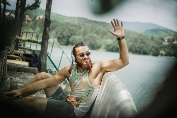 istock Young man with long hair and beard, attractive appearance with a surfboard on the beach next to a beautiful mountain lake. A tropical day next to here, a lake that has an environment similar to exotic beaches. 1314015111