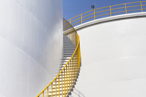 white fuel tanks against blue sky, white steel petroleum silo and white steel stair with yellow metal handrail.