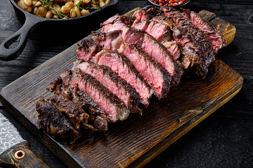 Rare rib eye steaks with herbs and spices set, on wooden serving board, with white beans and rosemary in cast iron pan, on black wooden table background