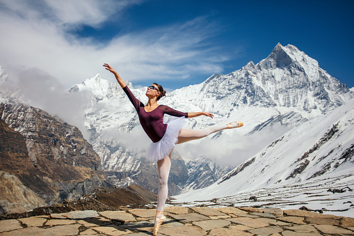 Young attractive ballerina performing ballet dance on Himalayas in Annapurna region with Machapuchare in background. Sve wearing white tutu, white stockings, purple leotard and ballet slippers.