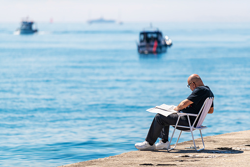 Istanbul, Turkey - April 22, 2021: One adult man sitting lounge chair and reading newspaper on lounge chair by the sea during the days of the epidemic in Istanbul, Turkey. Selective focus.