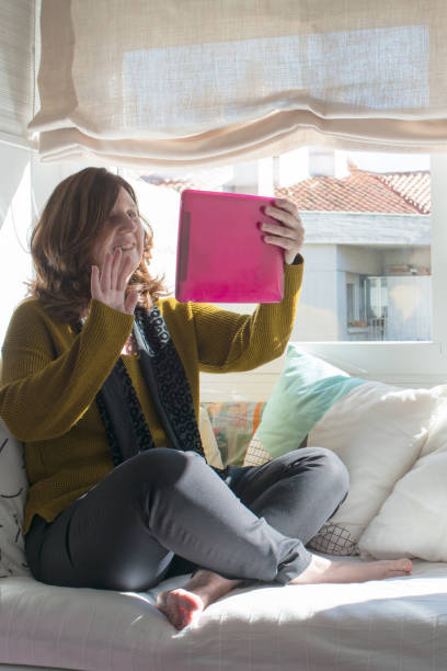 femme caucasienne âgée de 50 pendant un chat vidéo à la maison. cheveux bruns, assis sur un canapé d’une manière informelle, pieds nus, saluant le public. - women computer home interior brown hair photos et images de collection