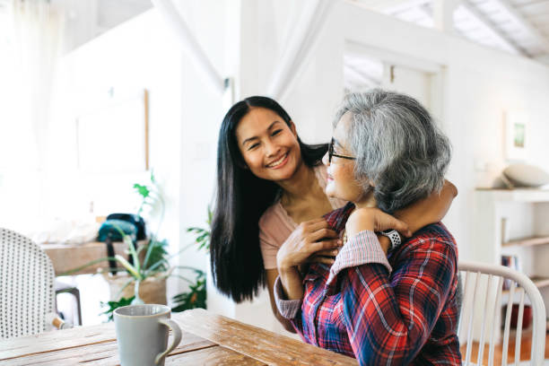 Loving adult daughter embracing cheerful senior mother at home Beautiful adult granddaughter hugging beautiful senior grandmother while sitting on the chair drinking coffee asian daughter stock pictures, royalty-free photos & images