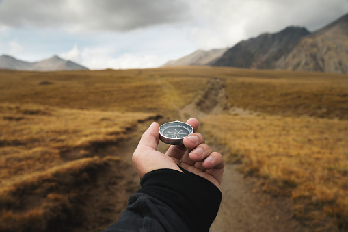 Man's hand holding a magnetic compass first-person view against the background of a high-altitude path and mountains.
