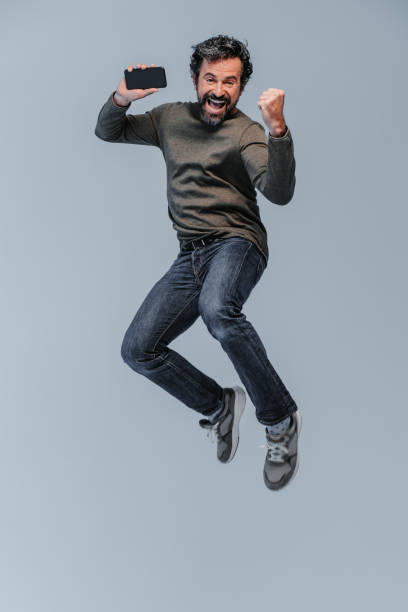 Studio shot of a exited happy screaming and exited mature man with casual clothings jumping high with his mobile phone in the hand stock photo