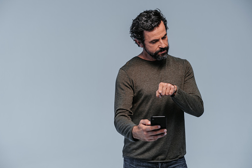 Studio shot of an impatient looking mature man with casual clothings and with his mobile phone one hand and pointing with the index finger to the phone with the other one standing on grey background. Part of a series with similar poses and different facial expressions.