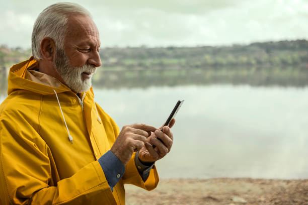 Senior man in woods on a raining day wearing yellow raincoat using smart phone