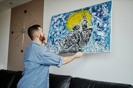 Man with beard hanging a painting on the wall at his living room