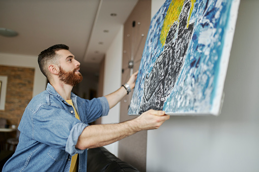 Man with beard hanging a painting on the wall at his living room