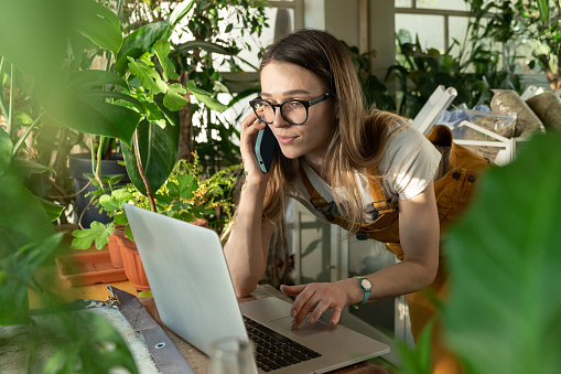 Freelance female work from home, woman gardener use laptop while talking with client on smartphone. Small floral business owner, florist entrepreneur make order online in home office at indoor garden