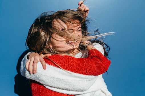 two friends, a blonde and a brunette, hugging each other in front of a blue wall on a windy day.