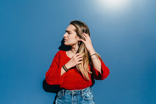young blonde in red sweater touching her hair in front of a blue wall in strong sunlight