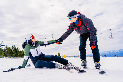 Young man helping a young female skier who fell down on mountain slope