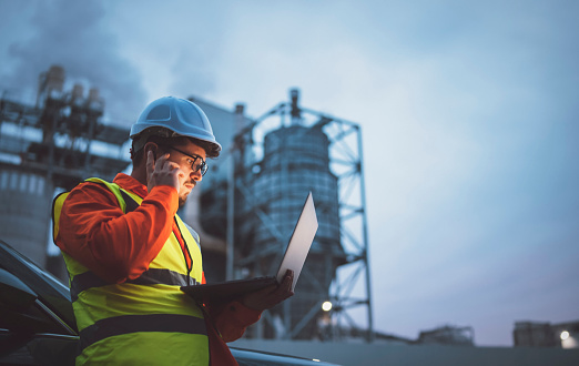 A shot of an young engineer wearing a helmet and using a laptop and hands free device during his night shirt in the oil rafinery. Engineering concept.