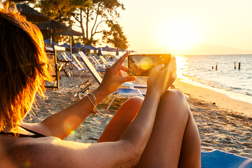 Summer vacation concept: A female person with sun kissed skin is taking a picture of a beautiful sunset with her mobile phone. She is sitting at the beach.\nCovid-19 travel ban
