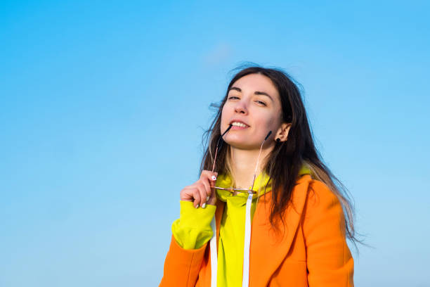 Good-looking carefree young woman. Pretty millennial lady wearing stylish vivid outfit having fun outdoors. Lifestyle portrait hipster girl holding sparkling sunglasses. Blue sky background.