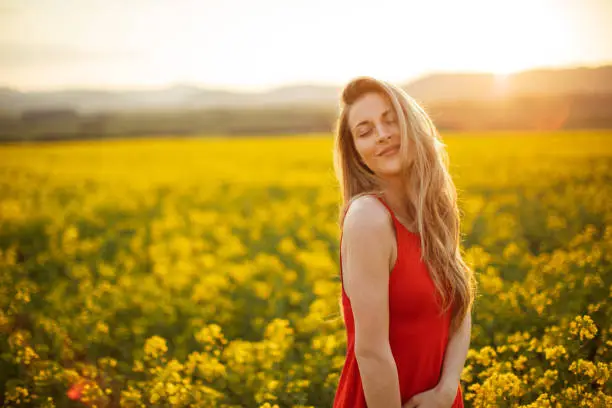 Beautiful young woman wearing bright red dress in yellow canola plant field. The field is spacious and the sun is showing in the background above the nearby hill. She is smiling and enjoying and celebrating freedom, happiness and positive thoughts and life.