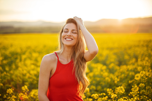 Beautiful young woman wearing bright red dress in yellow canola plant field. The field is spacious and the sun is showing in the background above the nearby hill. She is smiling and enjoying and celebrating freedom, happiness and positive thoughts and life.
