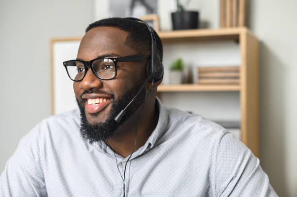 African-American office worker portrait Young and handsome African-American employee wearing glasses, headphones with a microphone, talking to customers, communicating, and providing customer service on a helpline at the call center call center stock pictures, royalty-free photos & images