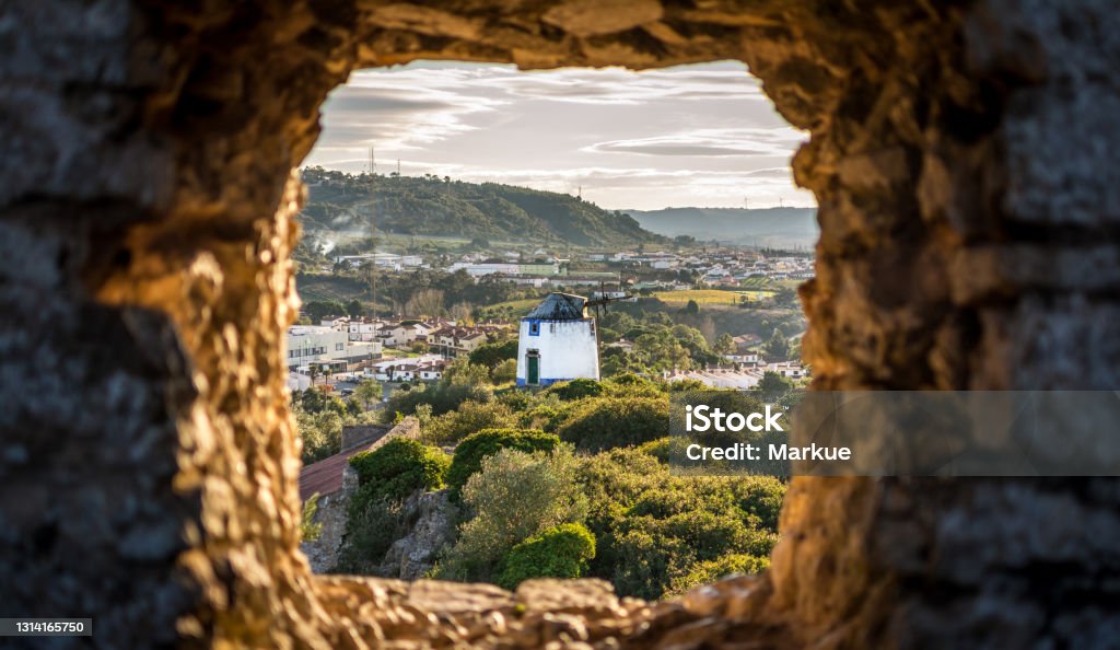 Old Windmill through Small Window in Obidos Fortress Wall, Portugal. Evening Sunlight Obidos Stock Photo