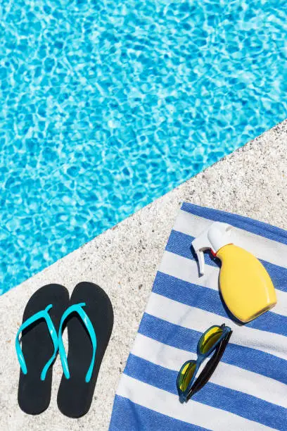Photo of Flip flops, sunscreen spray and sunglasses on a blue and white striped towel at the edge of a pool. Copy space.