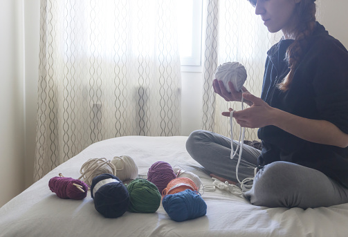 young white woman sitting on the bed with several balls of colored wool and cotton, holding a ball of white t-shirt yarn ball with her hands
