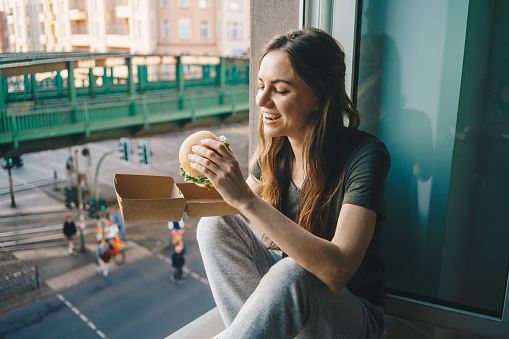 Woman eating take out burger at home in front of the open window