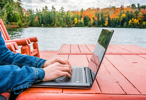 Young professional - digital nomad taking advantage of outdoors in autumn - working on laptop by the edge of a lake. Blurred background - autumn colors