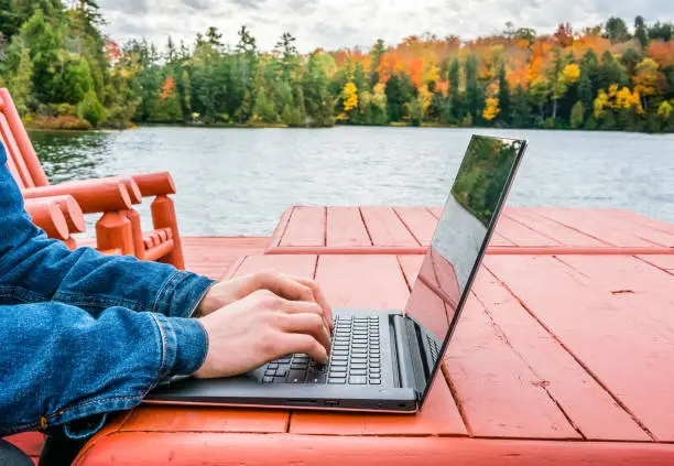 Photo of Adult male hands - digital nomad taking advantage of outdoors in autumn - working on laptop by the edge of a lake. Blurred background - autumn colors