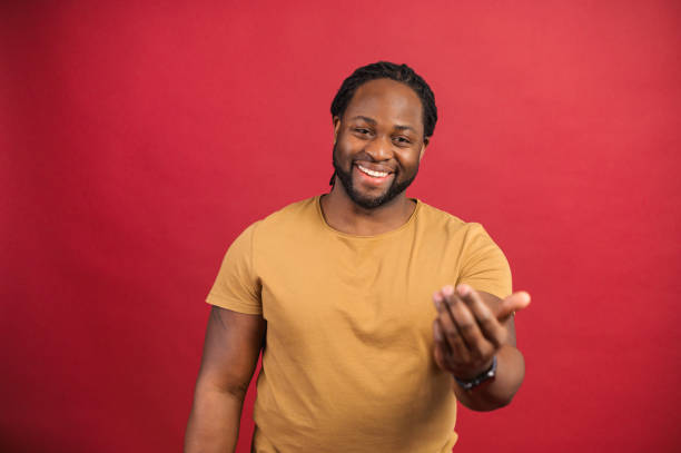 Cheerful African-American guy isolated on red Glad dark skinned man wearing casual shirt shows come on gesture with hands, cheerful African guy invites to come, isolated on red invitation gesture stock pictures, royalty-free photos & images