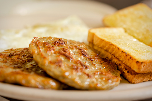 A dish at a breakfast diner including 2 pork sausage patties, two pieces of white toast, and 2 over easy eggs blurred in the background.  White plate.